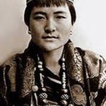 A black and white image of a bhutanese queen wearing a hat, earrings, and beaded necklace.