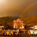 Tibetan style buildings with mountains and grey sky in the background. In the centre there is a rainbow.