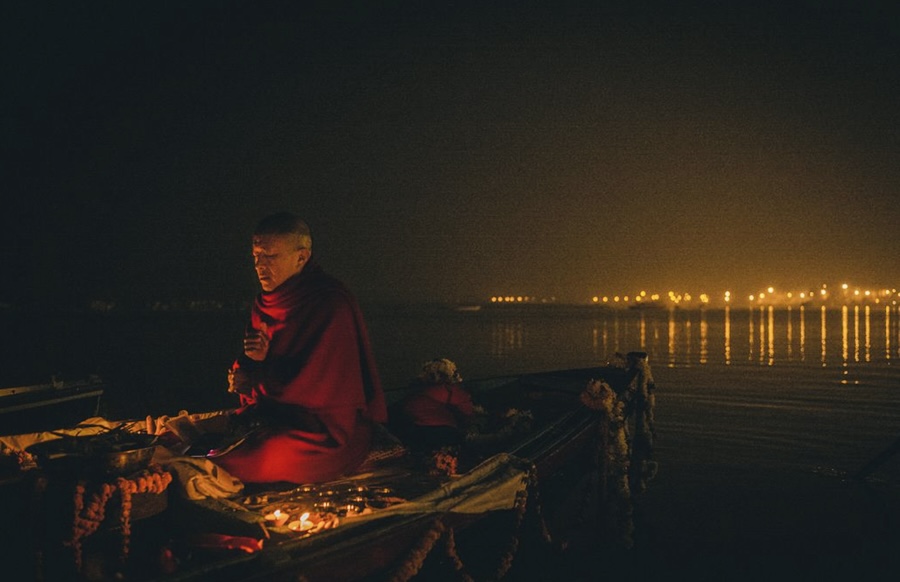 Dzongsar Khyenste Rinpoche is on a boat performing a fire puja in the nighttime, lights reflect off the water in the distance.