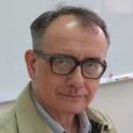 A man in heavy rimmed glasses wearing a tan jacket sits in front of a chalkboard.