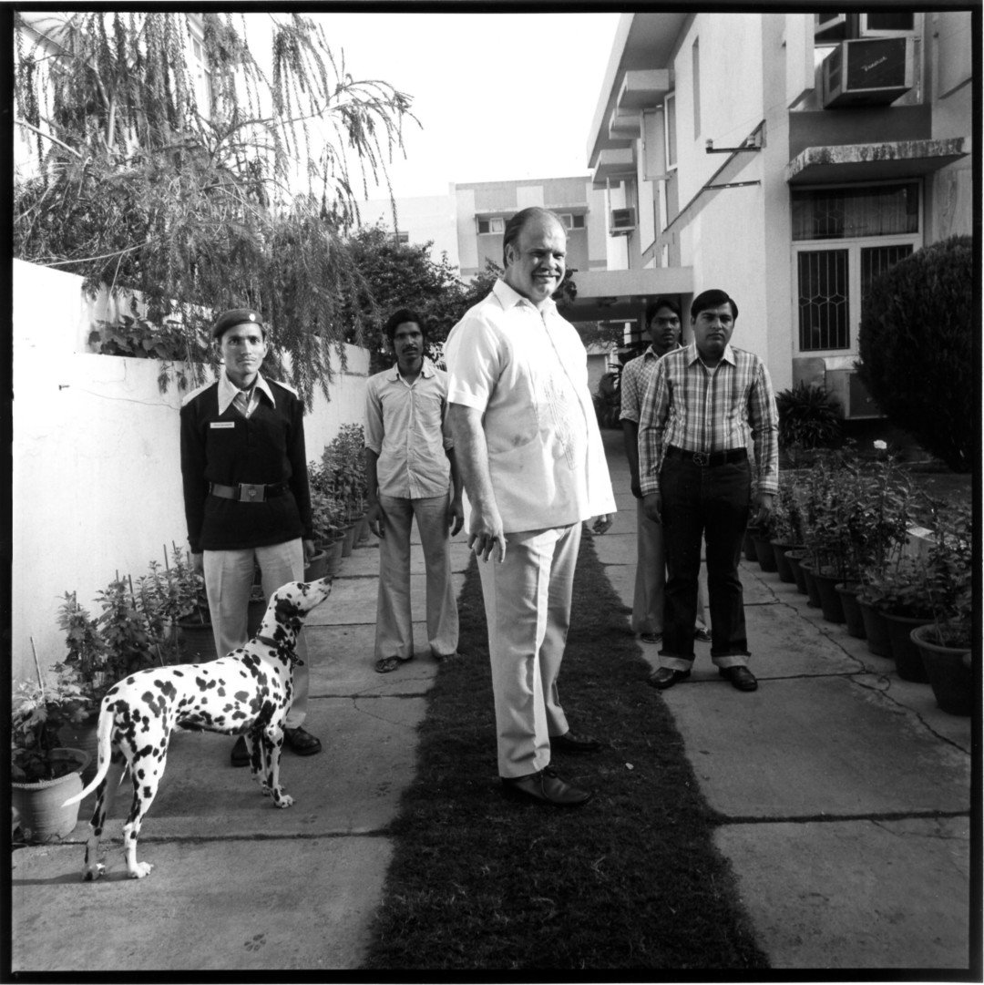 A gentle man stands in the center of a group of people with a spotted dog to his right.