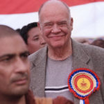 Gene Smith smiling in a crowd, wearing a big colorful ribbon on his lapel