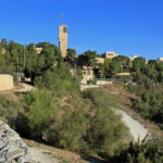 Many light brown buildings with a modern tower in the centre, a green lawn in front of it and a blue sky behind it.