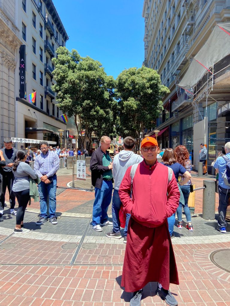 Standing in a San Francisco street with trees and buildings in the background is a monk wearing a red robe, a red jacket, and an orange cap.