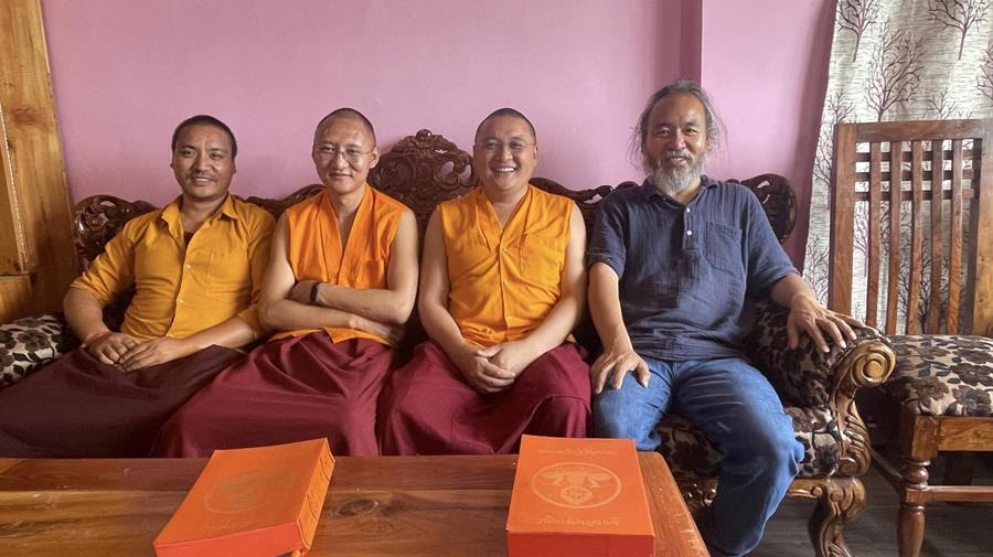 Three men in red and yellow robes and one with a blue shirt and jeans sitting on a sofa, smiling.