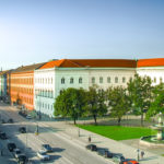 Old buildings with crimson roofs on both sides of a four-lane street. Fountains, green grass and trees on both sides.