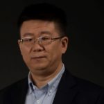 A man in a blue button down shirt and dark blazer wearing glasses sits for a portrait photo