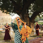 A woman smiling and holding marigold garlands. Red-robed monks behind her. The Sarnath stupa is in the background.