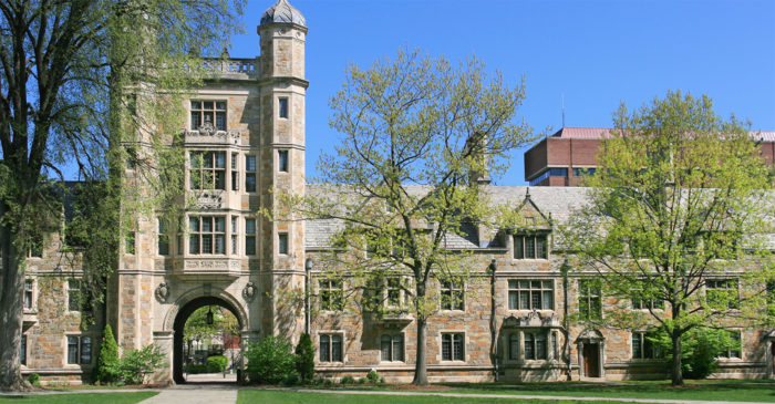Large old brown university building with green trees in front of it and blue sky in the background.